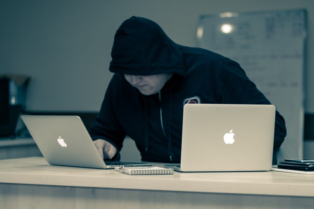 Un homme en sweat à capuche noir tape sur deux ordinateurs. Son visage est caché par la capuche.